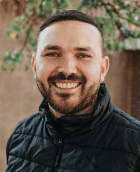 A smiling man with a beard wearing a black jacket, exuding a friendly and approachable demeanor.