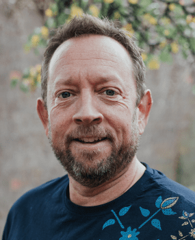 A man with a beard wearing a blue shirt, looking directly at the camera with a neutral expression.