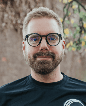 A man with glasses and a beard wearing a black t-shirt, looking directly at the camera with a neutral expression.